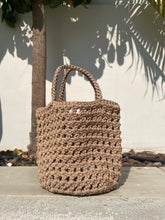Load image into Gallery viewer, Brown woven handbag on a stone surface with shadows and plants in the background
