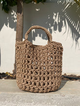 Load image into Gallery viewer, Brown woven handbag leaning against a light-colored wall with shadows cast by leaves.

