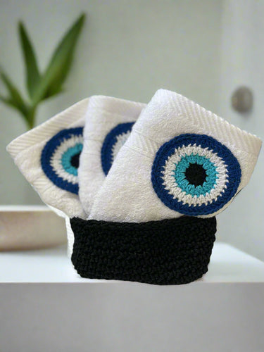 Three white towels with blue and black with an evil eye designs on a black holder against a white background