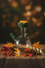 Load image into Gallery viewer, A clear glass bottle with a small side spout, topped with a dark green crocheted lid featuring a yellow-orange maple leaf and a matching green pom-pom charm, set on a light gray tiled floor.
