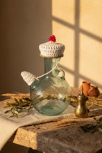 Load image into Gallery viewer, A clear glass bottle with a white crocheted cap and matching white neck cover, decorated with a small red flower on top, set on a light gray tiled floor.
