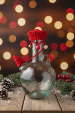 Load image into Gallery viewer, A clear glass bottle with a red crocheted hat and matching red cord with wooden beads, styled outdoors against a white wall and plant trunk.
