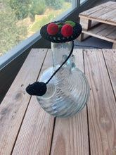 Load image into Gallery viewer, A clear ribbed glass bottle with a black crocheted cap featuring two red pom-poms and a black hanging loop, placed on a wooden table near a window.
