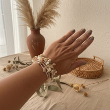 Load image into Gallery viewer, A close-up of a person's hand and wrist wearing a stack of natural shell bracelets, set against a bright outdoor swimming pool background.
