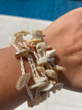 Load image into Gallery viewer, A close-up of a person's wrist wearing a stack of multi-strand bracelets made from natural cowrie shells on cream-colored cords, with a blue swimming pool in the background.
