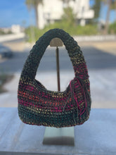 Load image into Gallery viewer, A crocheted shoulder bag with a multicolor pattern of black, teal, pink, and gold yarn, displayed on a metal stand against a blurred outdoor background.
