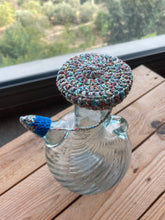 Load image into Gallery viewer, A clear glass bottle with a round, multicolored crocheted lid and a blue crocheted stopper, set on a wooden deck with a window and outdoor scenery in the background.
