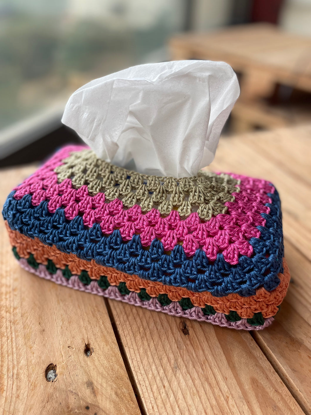 Colorful crocheted tissue box cover with tissues on a wooden surface