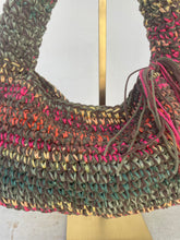 Load image into Gallery viewer, A close-up of a handmade woven shoulder bag made from multicolored ribbon yarn, featuring a crescent shape and hanging tassel details, displayed on a gold stand.
