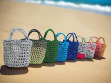 Load image into Gallery viewer, Row of colorful crochet bags on a sandy beach with ocean in the background
