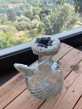 Load image into Gallery viewer, A clear ribbed glass bottle with a crocheted white and black neck cover, resting on a wooden table with a blurred outdoor garden background.
