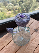 Load image into Gallery viewer, A handmade crocheted lavender and purple pet collar with a matching tag, displayed on the neck of a clear glass bottle on a wooden table with a window view of green trees in the background.
