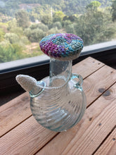 Load image into Gallery viewer, A clear glass bottle with a crocheted mushroom-shaped stopper in pastel purple, blue, green, and pink, resting on a wooden table with a window and outdoor landscape in the background.
