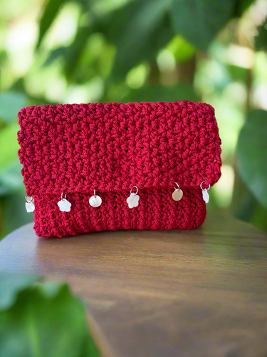 Red crochet clutch with silver charms on a wooden surface with green foliage in the background