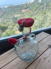 Load image into Gallery viewer, A clear ribbed glass bottle with side handles sits on a wooden table outdoors, decorated with a red crocheted mushroom cap and stem with a small gray felt mushroom.
