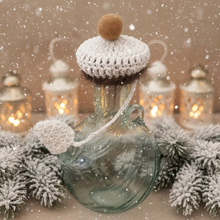 Load image into Gallery viewer, A clear glass bottle with a white crocheted cap and matching white hanging loop, topped with a small beige pom-pom, photographed on a light tiled floor.
