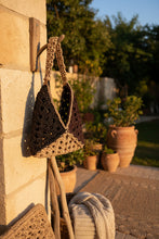 Load image into Gallery viewer, A handmade crocheted shoulder bag with a dark brown triangular body, a beige textured strap, and a black woven backing, displayed on a reflective white surface with dried palm leaves in the background.
