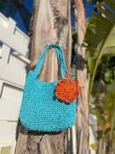 Load image into Gallery viewer, Blue crocheted bag with an orange flower attachment hanging on a wooden post.
