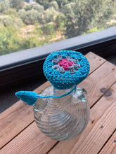 Load image into Gallery viewer, A turquoise crocheted wine bottle stopper with a pink and gray floral center and a matching turquoise loop, shown resting on a clear glass bottle on a wooden table with a window and outdoor scenery in the background.
