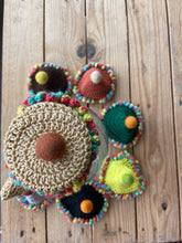 Load image into Gallery viewer, A handmade crochet teapot cover set with colorful pom-pom trim and felted tops, arranged on a rustic wooden surface. The set includes multiple covers in different colors: beige, green, yellow, black, brown, and red.
