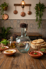 Load image into Gallery viewer, A clear glass bottle with a crocheted dark brown cap and a small white felt acrond on a black cord on a table with Lebanese food 
