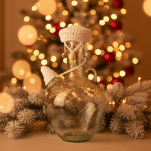 Load image into Gallery viewer, A clear glass bottle with a rounded body, small handle, and narrow neck, decorated with a white crocheted cap and matching white cord tied around the neck. The bottle is photographed outdoors on a stone surface with dried plant trunks and mulch in the background.
