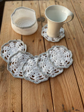 Load image into Gallery viewer, Set of crocheted coasters on a wooden surface with a cup and small container.

