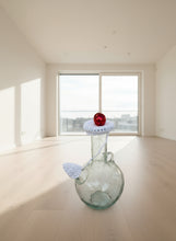 Load image into Gallery viewer, Small glass bottle with a red ball on top, white string, and crocheted lid against a tiled wall background.
