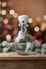 Load image into Gallery viewer, A clear glass bottle with a white crocheted cap and small hanging beads, photographed outdoors against a white wall and plant trunk.
