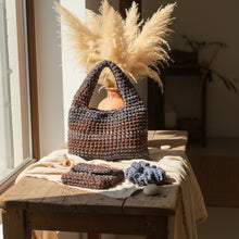 Load image into Gallery viewer, A handmade crocheted tote bag in variegated brown and blue brown gray yarn, displayed outdoors next to a tree trunk with a matching crocheted pouch and scrunchie on a concrete surface.
