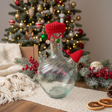 Load image into Gallery viewer, A clear glass bottle with a textured finish, decorated with a bright red crocheted Santa hat and matching red yarn beard, set outdoors on a stone surface.
